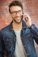 Smiling trendy guy with eyeglasses standing by brick wall talking on phone