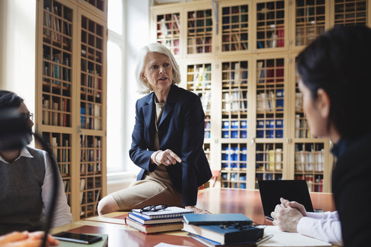 Senior professional gesturing while sitting on table during meeting in library - Powered by Adobe