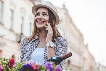 Inviting her friends to join her in the park. Beautiful young woman talking on the mobile phone and smiling while standing near her vintage bicycle with basket of flowers.