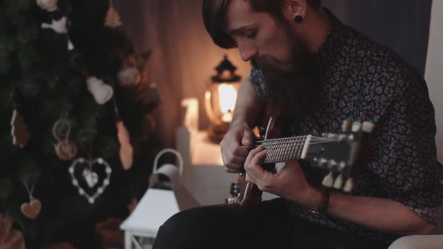 Hipster with beard, moustache and ear plugs playing electric guitar at New Year's Eve with Christmas tree on background.