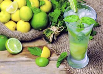Fresh glass of lemon soda with ice and mint placed with set of fresh mint and jute bag of organic lemon on rustic wooden table.