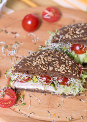 Very healthy breakfast full of vitamins. . Meal of whole wheat dark/brown rye bread, curd cream cheese, radish, alfalfa sprouts, ham, cherry tomatoes, green salad on wooden cutting board. Closeup