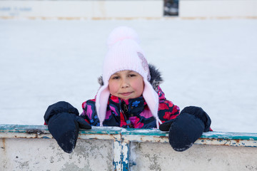 Young girl with red frozen cheeks standing near the board of courtyard ice rink while skating