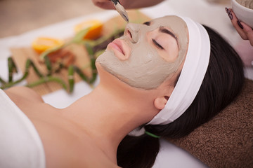Beautiful young woman receiving gray facial mask in the beauty salon