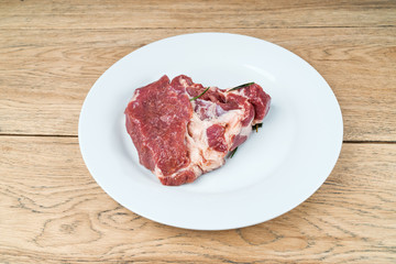 Big piece raw meat with rosemary leaves lay on a white plate on old wooden table
