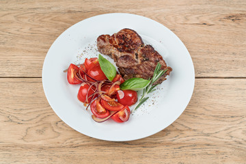 Pork steak on the bone with tomatoes and onions served on a white plate on old wooden table