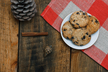 Chocolate cookies in a white plate on a checkered tablecloth on wooden background. Near cones and cinnamon stick, roughly. View from above