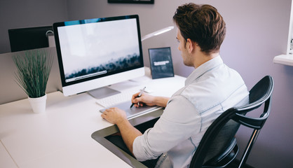 Young worker sitting in an office at the computer. Freelancer in a blue shirt. The designer sits in front of window in the workplace.