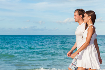 Fototapeta na wymiar Holiday couple relaxing walking on beach at sunset on honeymoon travel vacation holidays. Young interracial couple in love wearing white dress and shirt, Asian woman and Caucasian man.