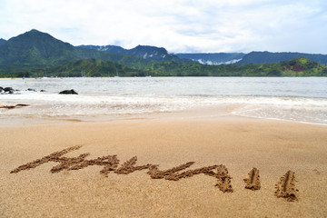 Hawaii word written on sand on hawaiian beach. Handwriting text texture on holiday background. Famous destination Hanalei bay in Kauai, USA with ocean and mountains.