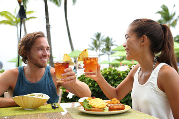 Happy multiracial couple toasting cheers with alcoholic hawaiian drinks, mai tai, Hawaii experience. Summer travel holidays, people enjoying local food meal at outdoor terrace restaurant of resort.