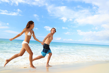 Happy beach couple vacation getaway. Young people in bikini and swimwear running holding hands together having fun on tropical beach paradise. Perfect blue ocean water and white sand.