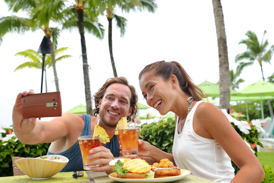 Happy young interracial couple on summer holiday going out for dinner enjoying local food meal at outdoor terrace restaurant taking a selfie picture with mobile phone for vacation memories.