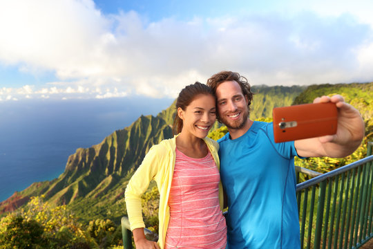 Happy interracial travel couple taking selfie mobile phone picture at Kalalau lookout on na pali coast viewpoint in Kauai, Hawaii with amazing scenic landscape. Famous popular hawaiian attraction.