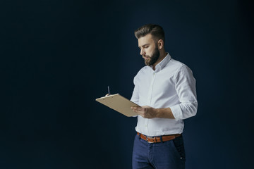 Young bearded businessman, dressed in a white shirt and blue pants, standing and making notes on a clipboard, placed in his hands. In the background dark blue wall. The man wrote in pencil.