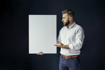 Young bearded businessman, dressed in a white shirt standing and holding a blank white poster. In...