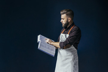 Side view. A young bearded man, dressed in a dark brown shirt and bright apron, holding a thick book with recipes and reading. In the background dark blue wall.