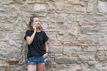 Front view of a young woman dressed in denim shorts and a black t-shirt, standing and talking on the phone. In the background is bright stone wall. Girl uses a digital gadget.