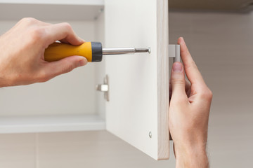The worker sets a new handle on the white cabinet with a screwdriver - obrazy, fototapety, plakaty