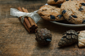Chocolate chip cookies on rustic background. Nearby lie cinnamon sticks and cones. Behind is tree with toys and garlands. New Year holiday.