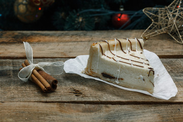 Beautiful homemade cake cheesecake on a white paper on the wooden background with cinnamon stick. Near Christmas gifts, Christmas tree and toys. Conceived to celebrate the new year.