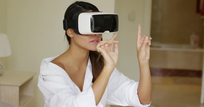 Young woman wearing virtual reality glasses gesturing with her hands as she interacts with her simulated environment while relaxing on her bed