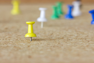Yellow pushpin surrounded by white, green a blue pushpins in a cork board.