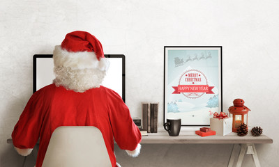 Santa Claus planning trip on computer. Gifts, books, tea, picture and lantern on table.
