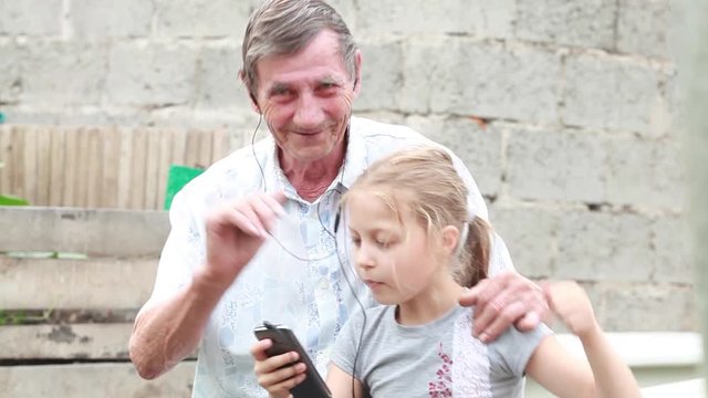 Grandfather and granddaughter listening to music on headphones and dancing holding a cell phone in the park