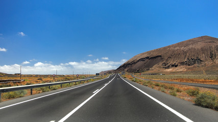 Asphalt road disappearing over the horizon through volcano mountain hillsides. White clouds on a blue sky. Lanzarote, Canary Islands, Spain