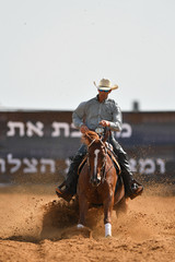 The front view of a rider in cowboy chaps, boots and hat on a horseback running ahead and stopping the horse in the dust.