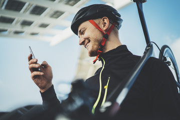 Young happy smiling cyclist man using smartphone while resting after workout, road bicycle on arms, cheerful sportsman checking fitness results via cellphone while riding a bike at the sunny morning