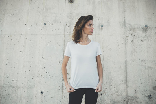 Young hipster girl wearing blank white t-shirt, stone wall background with copy space for content or design, mockup of template t-shirt, attractive woman standing against concrete wall background