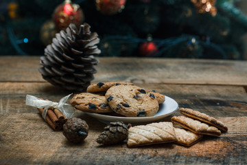 Fototapeta na wymiar Chocolate chip cookies on rustic background. Nearby lie cinnamon sticks and cones. Behind is tree with toys and garlands. New Year holiday.
