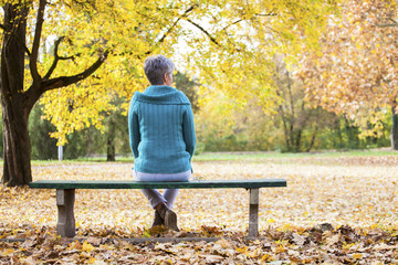 Older women with gray hair sits alone on a bench in autumn park and grieves. Sad and depressing scenes of the old lady in the natural environment.