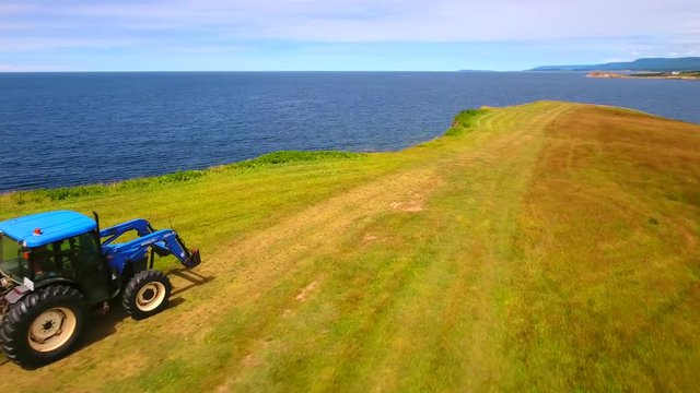 Aerial rotating shot as farmer cuts grass on field by ocean cove