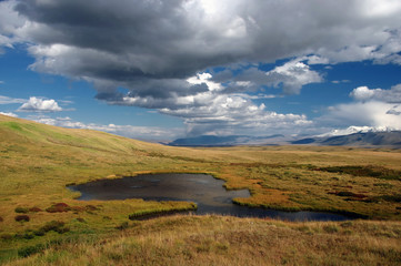 Alpine steppe meadows with yellow and green grass on a plateau with small lake on the background of mountains with snow and ice under a blue sky and dramatic clouds Ukok Altai mountains Siberia Russia