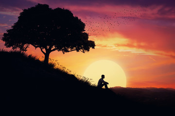 Fototapeta premium Boy sit alone on a hill in the center of nature, over a sunset background. Standing away from the crowd, waiting for the healing power of the nature.