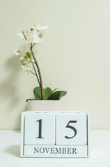 Closeup white wooden calendar with black 15 november word with white orchid flower on white wood desk and cream color wallpaper in room textured background , selective focus at the calendar