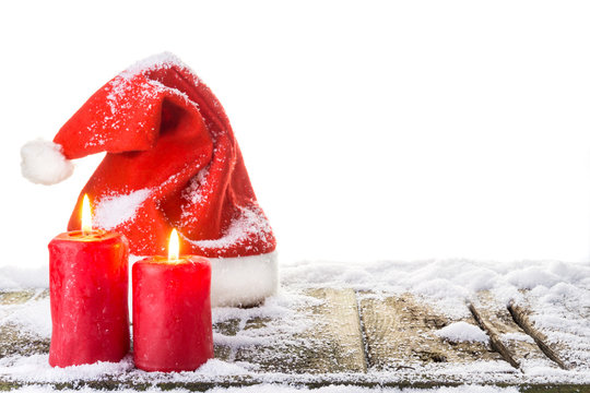 Weihnachtsmütze und 2 brennende Kerzen auf verschneitem Holzboden vor reinweißem Hintergrund