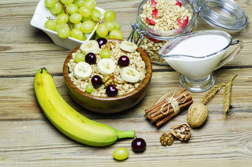 Breakfast with muesli and granola in wooden bowl placed with fresh fruits, cinnamon, nuts and jug of warm milk on wooden background.