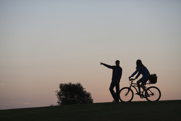 Obraz premium Silhouette of a couple walking through a park in San Diego at dusk. The woman is riding a bike and the man is walking. 