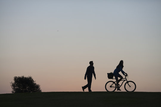 Silhouette of a couple walking through a park in San Diego at dusk. The woman is riding a bike and the man is walking. 