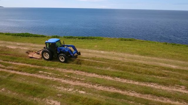 Aerial rotating shot as farmer cuts the grass on field by ocean cove