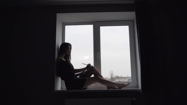 Attractive young brunette girl sitting on the windowsill and writing in notebook