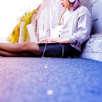 young beautiful caucasian purple grey hair woman outdoor in the city sitting on the floor listening music with head phones and computer leaning on her knee - technology, music, social network concept