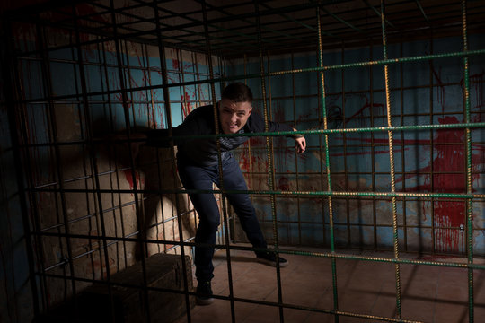 Man imprisoned in a metal cage with a blood splattered wall behi