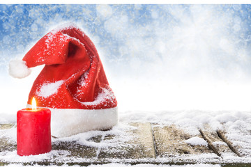 Weihnachtsmütze und brennende Kerze auf verschneitem Holzboden vor blauem Hintergrund mit Bokeh...