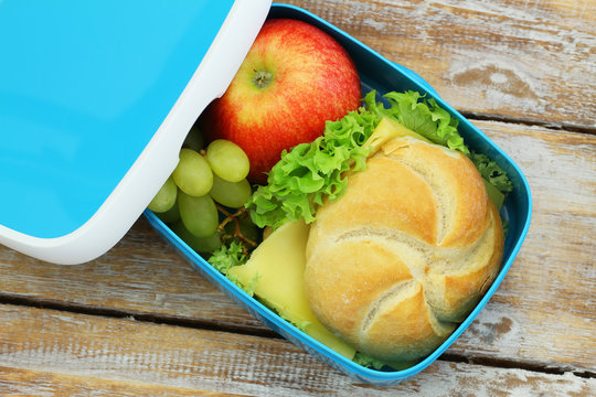 Open lunch box containing bread roll with cheese and lettuce, red apple and grapes on rustic wood
