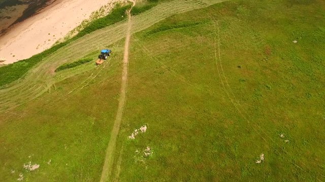 Aerial shot as a farmer cuts grass on a field by ocean cove